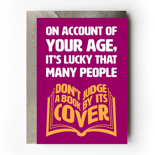 Text on card is white and yellow against a burgundy background and states "On account of your age, it's lucky that many people don't judge a book by its cover"  The 'don't judge a book by its cover' text is in the shape of an open book. Card measures 105 x 150mm and is supplied with a very striking pewter-effect eco-Kraft envelope.
