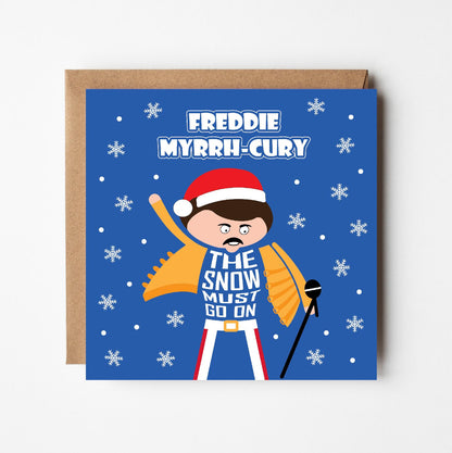Christmas card with Freddie Mercury wearing Santa hat with pun text saying Freddie MyrrhCury and the Snow must go on. Printed on blue background with snowflakes.